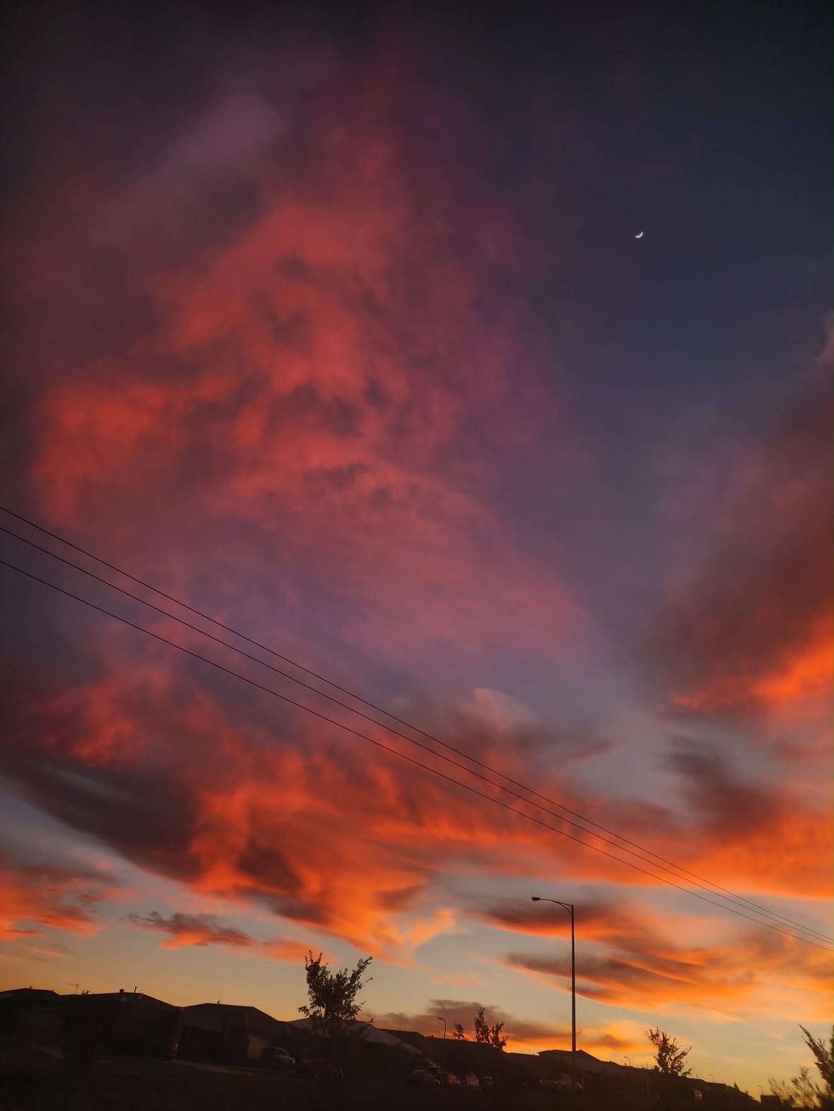 A crescent moon hangs in a darkening sky as clouds burn orange and pink above power lines and rooftops.