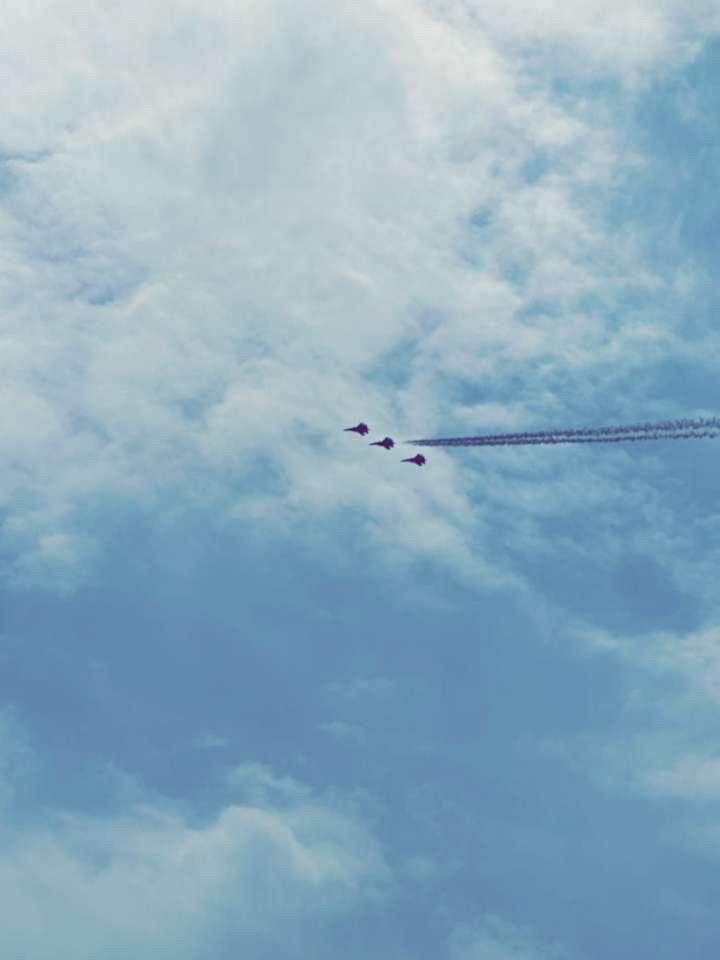 Three jets trailing dark smoke cut across a pale blue sky layered with soft clouds.