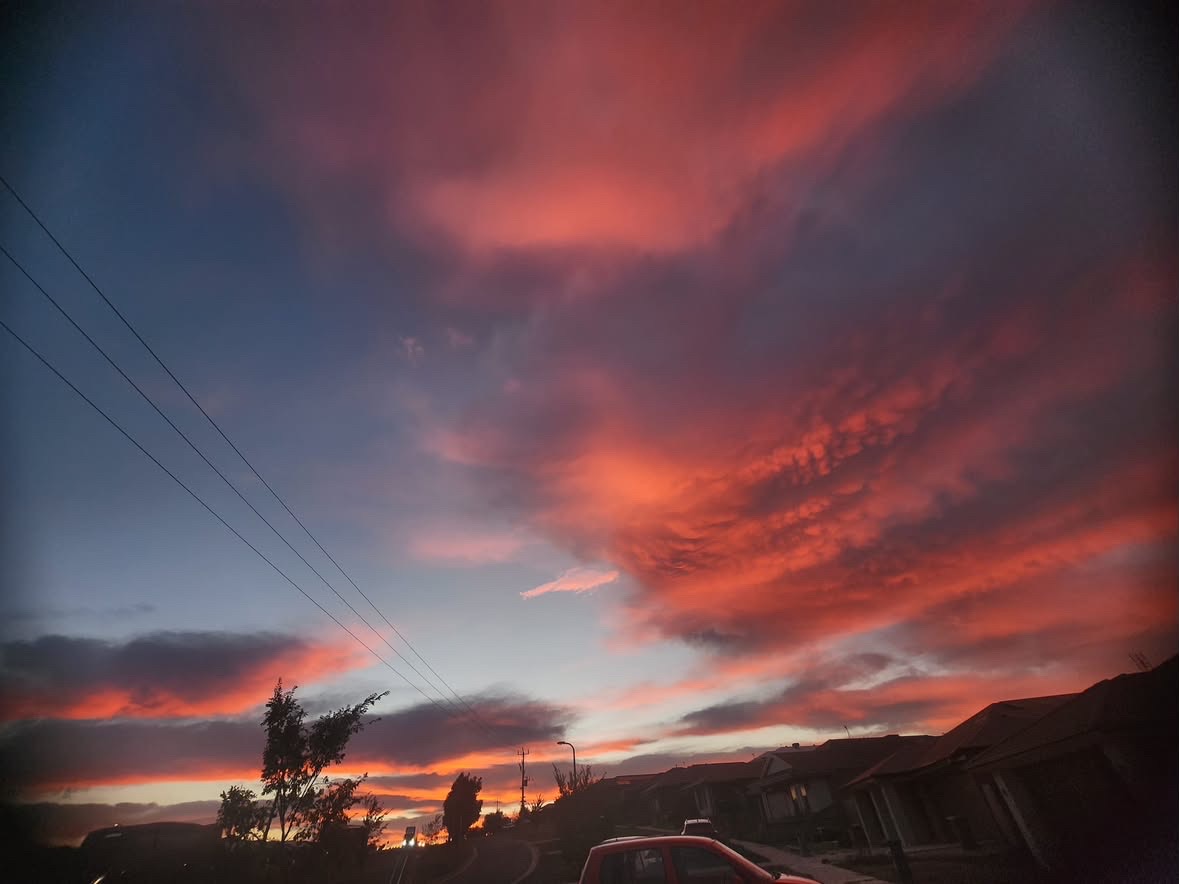 A sweep of red and orange cloud stretches over a suburban street at sunset, a car parked below.